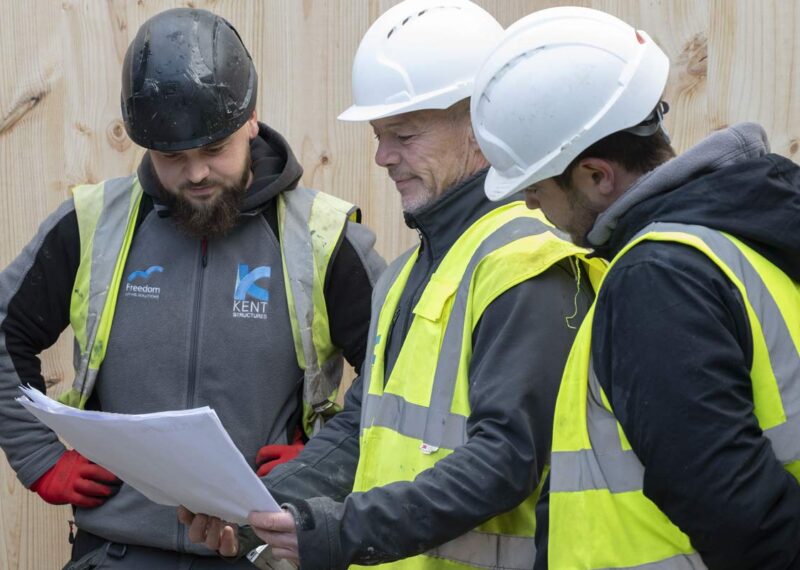 Architect and site workers in hard hats on a construction site reviewing value engineered plan.